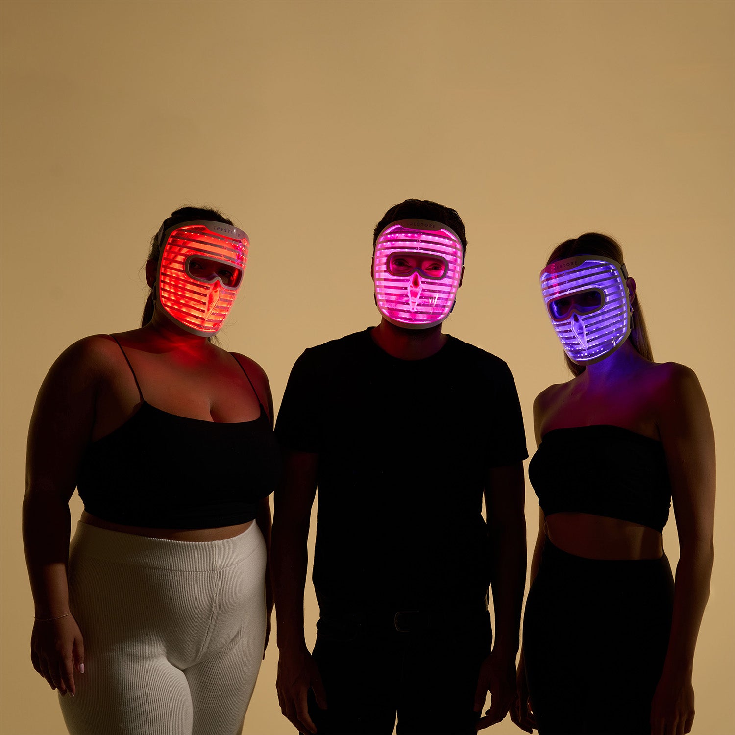 Man standing between two women, each wearing the iRESTORE LED face mask with the mask glowing in red, purple, and blue light, showcasing the different light therapy modes.