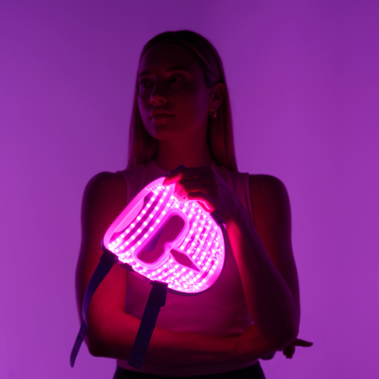 Woman holding the iRESTORE LED face mask, with the mask glowing in purple light, against a slightly dark background.