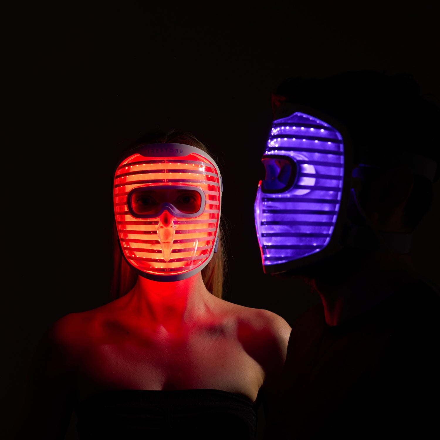 Front view of a woman wearing the iRESTORE LED face mask glowing red, standing beside a man in side view wearing the iRESTORE LED face mask glowing blue.