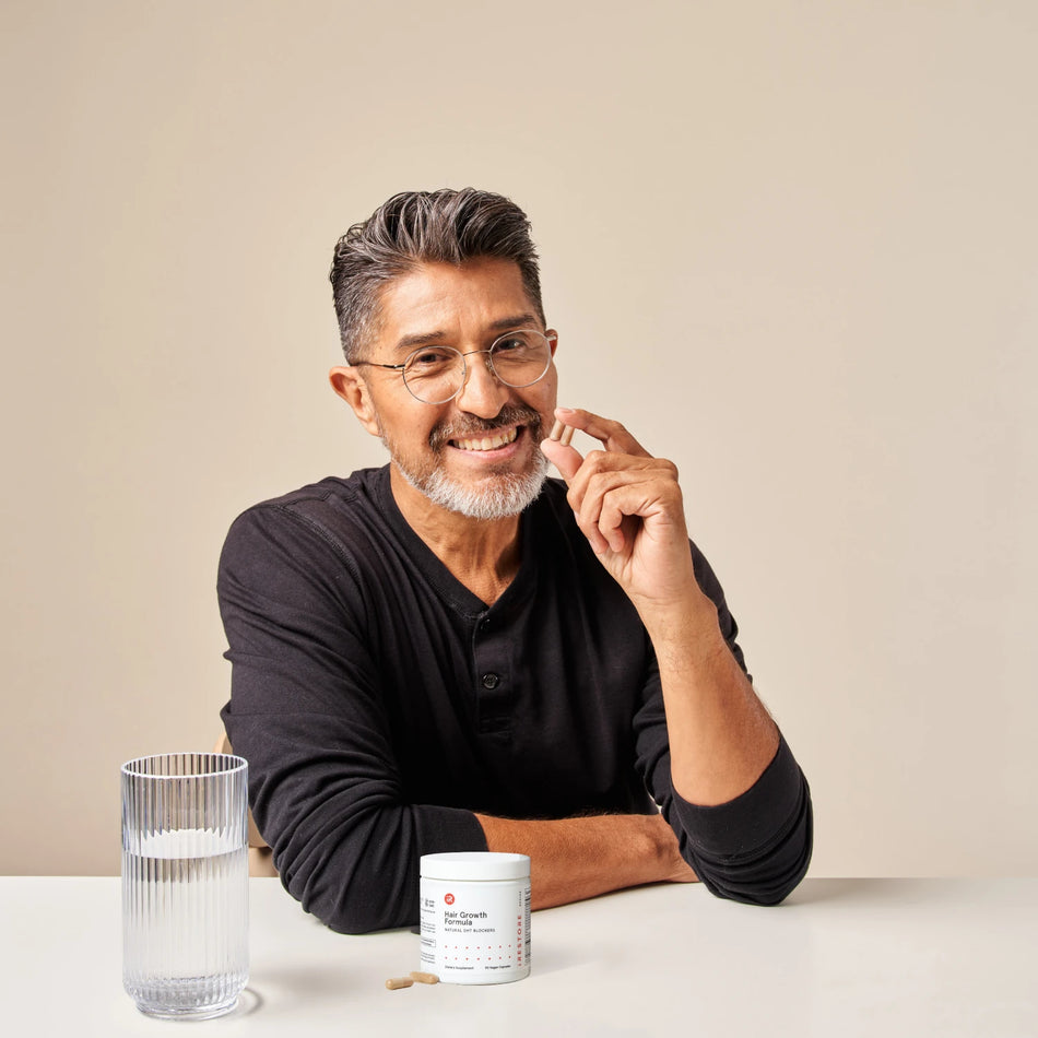 Image of a sitting man with Hair Growth Formula tablet in his hand. In front of him is a table with the cylindrical jar of the supplement placed on top of the table with a glass of water beside it.